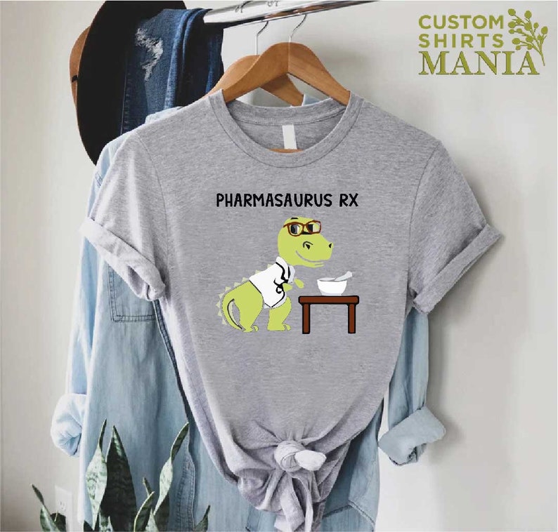 May include: A grey t-shirt featuring a cartoon dinosaur wearing glasses and a lab coat. The dinosaur is holding a mortar and pestle. The text "PHARMASAURUS RX" is printed on the shirt.