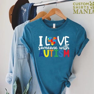 K&ouml;nnte beinhalten: Tealblaues T-Shirt mit einer wei&szlig;en Grafik, auf der "Ich liebe jemanden mit AUTISMUS" steht. Die Grafik enth&auml;lt ein Herz aus Puzzle-Teilen in Rot, Blau, Gelb und Gr&uuml;n.