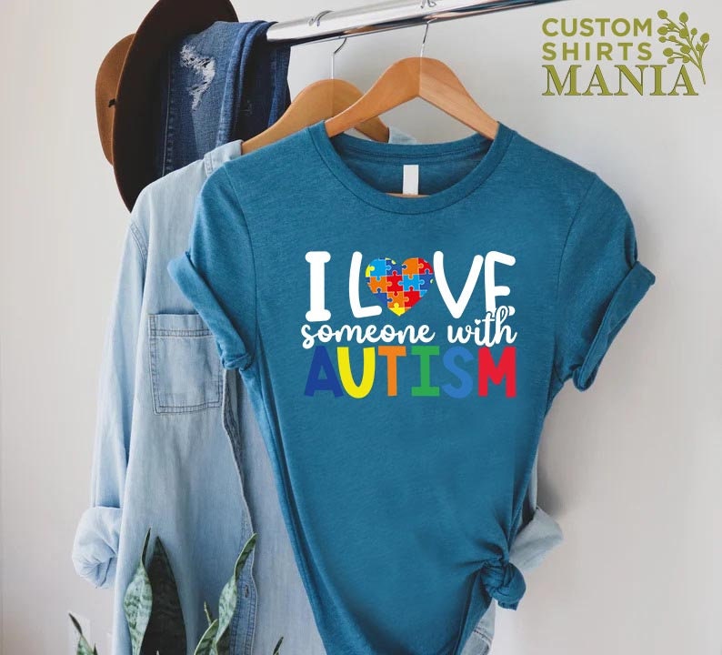 K&ouml;nnte beinhalten: Tealblaues T-Shirt mit einer wei&szlig;en Grafik, auf der "Ich liebe jemanden mit AUTISMUS" steht. Die Grafik enth&auml;lt ein Herz aus Puzzle-Teilen in Rot, Blau, Gelb und Gr&uuml;n.