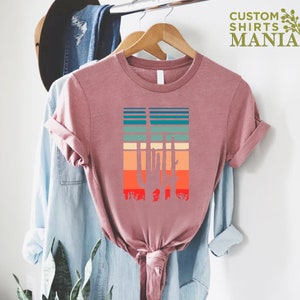May include: A pink t-shirt with a retro-style graphic of a cactus silhouette in front of a sunset. The graphic features a variety of colors, including orange, red, yellow, green, and blue.
