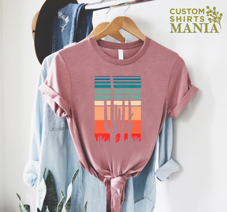 May include: A pink t-shirt with a retro-style graphic of a cactus silhouette in front of a sunset. The graphic features a variety of colors, including orange, red, yellow, green, and blue.