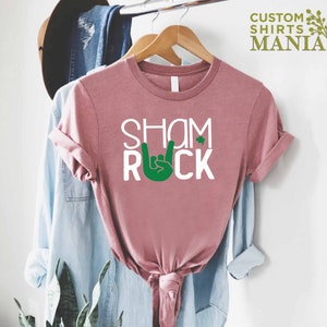 Peut inclure: Un t-shirt rose avec le texte "Shamrock" et une main verte faisant un signe de rock and roll. Le mot "Rock" est sous la main.