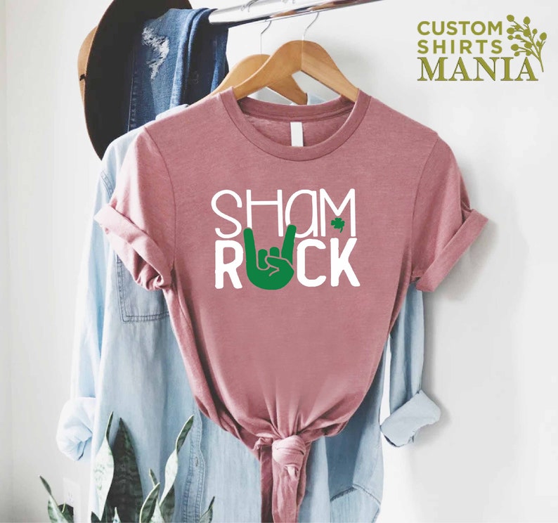 Peut inclure: Un t-shirt rose avec le texte "Shamrock" et une main verte faisant un signe de rock and roll. Le mot "Rock" est sous la main.