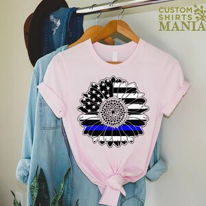 May include: A pink t-shirt featuring a black and white sunflower design. The sunflower has a blue line running through the centre, representing the thin blue line.