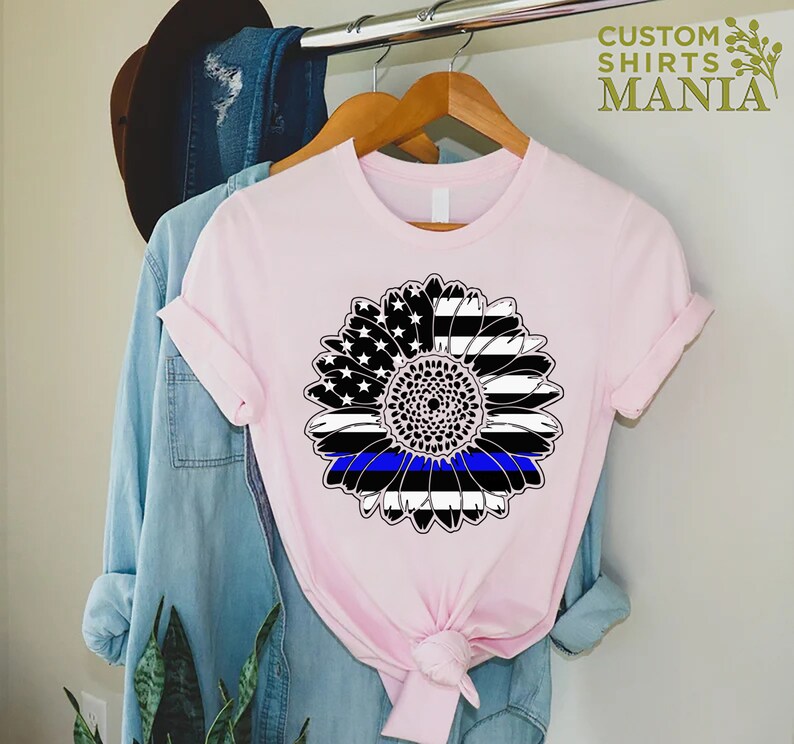 May include: A pink t-shirt featuring a black and white sunflower design. The sunflower has a blue line running through the centre, representing the thin blue line.