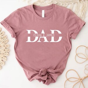 Pu&ograve; includere: Una maglietta color rosa polvere con la parola "DAD" in lettere bianche a blocchi. Il testo "Olivia & James" &egrave; scritto in lettere bianche pi&ugrave; piccole sotto la parola "DAD".