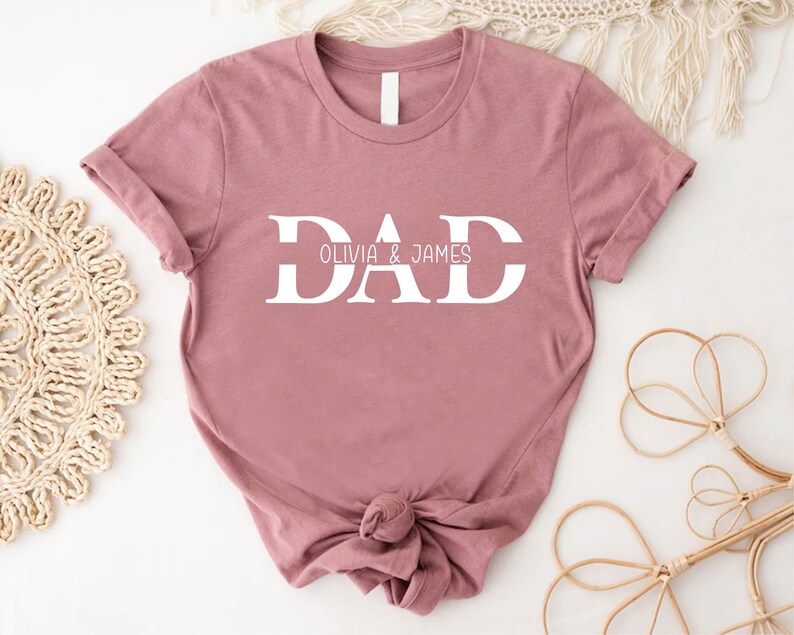 Pu&ograve; includere: Una maglietta color rosa polvere con la parola "DAD" in lettere bianche a blocchi. Il testo "Olivia & James" &egrave; scritto in lettere bianche pi&ugrave; piccole sotto la parola "DAD".
