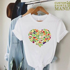 May include: White t-shirt featuring a heart-shaped design composed of various colorful vegetables. The shirt is hanging on a wooden hanger. The text "CUSTOM SHIRTS MANIA" is printed in green on the upper right.