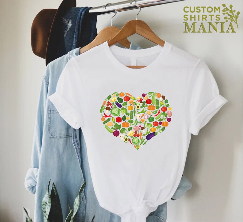 May include: White t-shirt featuring a heart-shaped design composed of various colorful vegetables. The shirt is hanging on a wooden hanger. The text "CUSTOM SHIRTS MANIA" is printed in green on the upper right.