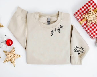 May include: A beige sweatshirt with the word "gigi" in black script on the chest. The right sleeve has the names "Emily Robert" and a heart. Christmas decorations, including a wrapped gift and star ornaments, are arranged around the sweatshirt.