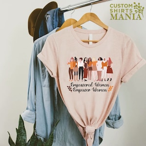 May include: A pink t-shirt featuring a graphic of a group of diverse women standing together. The text "Empowered Women Empower Women" is printed below the graphic.