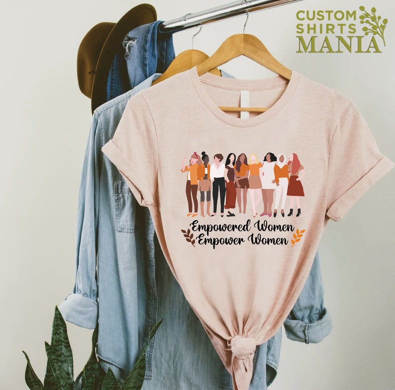 May include: A pink t-shirt featuring a graphic of a group of diverse women standing together. The text "Empowered Women Empower Women" is printed below the graphic.