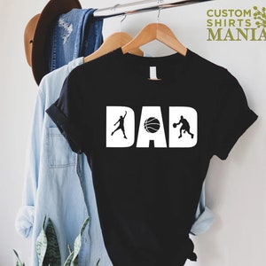 Op de afbeelding: Zwart T-shirt met witte tekst die "DAD" zegt met een basketbal en twee basketballers aan weerszijden van het woord.