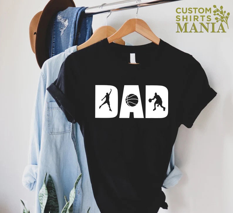 Op de afbeelding: Zwart T-shirt met witte tekst die "DAD" zegt met een basketbal en twee basketballers aan weerszijden van het woord.