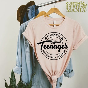 May include: A pink t-shirt with a black circle graphic that says "Warning Official Teenager May Contain Attitude".