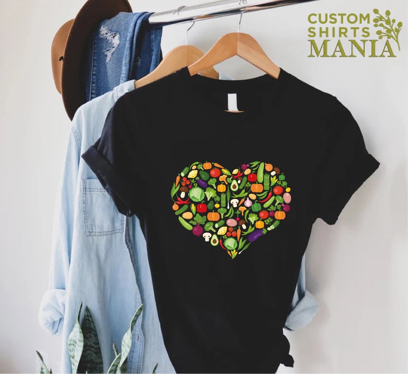 May include: Black t-shirt featuring a heart-shaped design filled with colorful vegetables. The vegetables include tomatoes, peppers, avocados, and more. The text "CUSTOM SHIRTS MANIA" is printed in green on the upper right.
