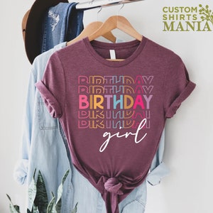 May include: A maroon t-shirt with the text "BIRTHDAY girl" in rainbow colours. The text is stacked vertically with the word "BIRTHDAY" in a larger font.
