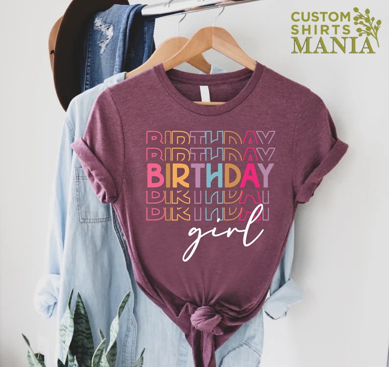 May include: A maroon t-shirt with the text "BIRTHDAY girl" in rainbow colours. The text is stacked vertically with the word "BIRTHDAY" in a larger font.