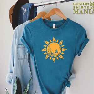 Puede incluir: Una camiseta azul turquesa con un gr&aacute;fico de sol amarillo que presenta una mariposa y detalles florales. El texto "CUSTOM SHIRTS MANIA" est&aacute; en la esquina superior derecha de la imagen.