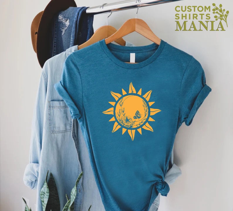 Puede incluir: Una camiseta azul turquesa con un gr&aacute;fico de sol amarillo que presenta una mariposa y detalles florales. El texto "CUSTOM SHIRTS MANIA" est&aacute; en la esquina superior derecha de la imagen.