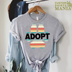 May include: Gray t-shirt with a colorful paw print design and the word "ADOPT" in black text.