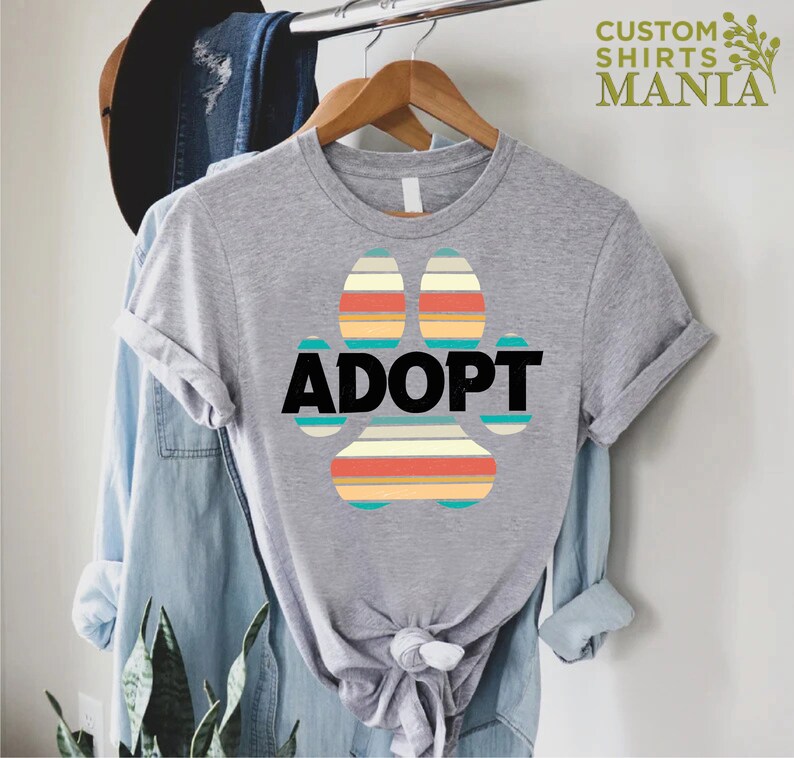 May include: Gray t-shirt with a colorful paw print design and the word "ADOPT" in black text.