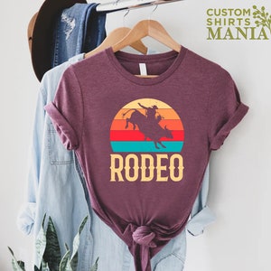 May include: A maroon t-shirt with a vintage-style sunset graphic featuring a silhouette of a cowboy riding a bull. The text "RODEO" is printed below the graphic.