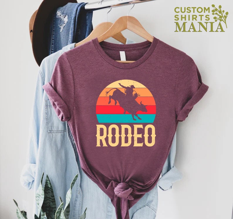 May include: A maroon t-shirt with a vintage-style sunset graphic featuring a silhouette of a cowboy riding a bull. The text "RODEO" is printed below the graphic.