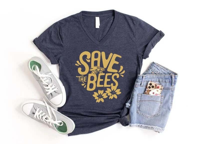 May include: A navy blue V-neck t-shirt with the text "SAVE THE BEES" in gold. The design includes a bee and flowers. The shirt is paired with gray sneakers and denim shorts.
