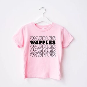 Może przedstawiać: R&oacute;żowa koszulka z kr&oacute;tkim rękawem z napisem "WAFFLES" powt&oacute;rzonym pięć razy czarnym tekstem.