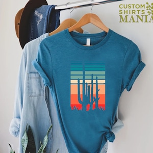 May include: A teal blue t-shirt with a retro-style graphic of a cactus silhouette against a colorful striped background. The shirt is hanging on a hanger.