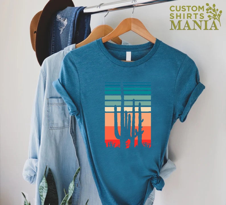 May include: A teal blue t-shirt with a retro-style graphic of a cactus silhouette against a colorful striped background. The shirt is hanging on a hanger.