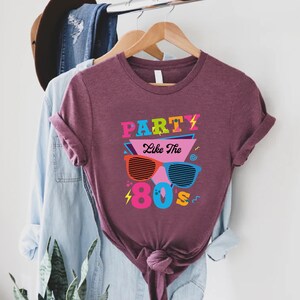 K&ouml;nnte beinhalten: Ein maroonfarbenes T-Shirt mit dem Text "Party Like The 80s" in bunten Buchstaben und einer Grafik von Retro-Sonnenbrillen.