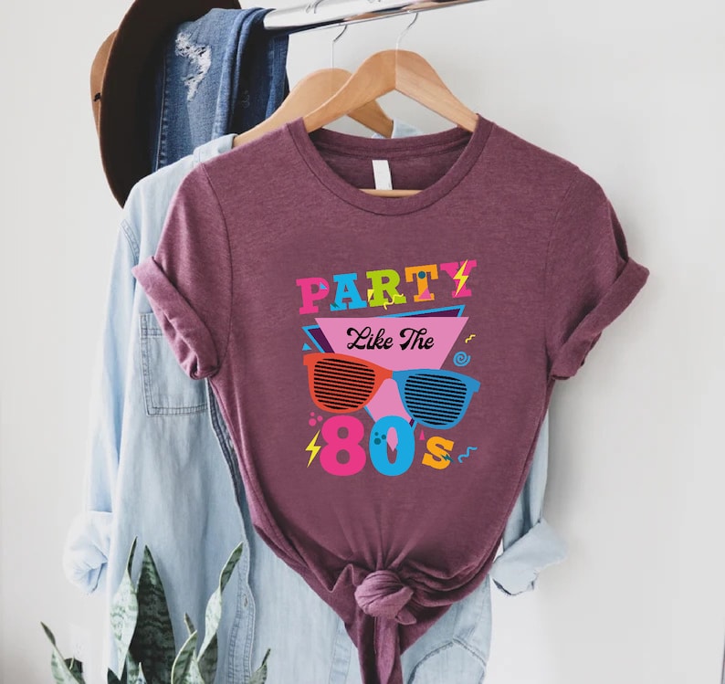K&ouml;nnte beinhalten: Ein maroonfarbenes T-Shirt mit dem Text "Party Like The 80s" in bunten Buchstaben und einer Grafik von Retro-Sonnenbrillen.