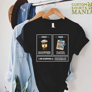 May include: Black t-shirt with a white graphic design. The graphic shows two boxes, one with a cup of coffee and the text "First - Coffee" and the other with a clipboard and pen and the text "Then - Data". Below the boxes is the text "I am earning a: Break".