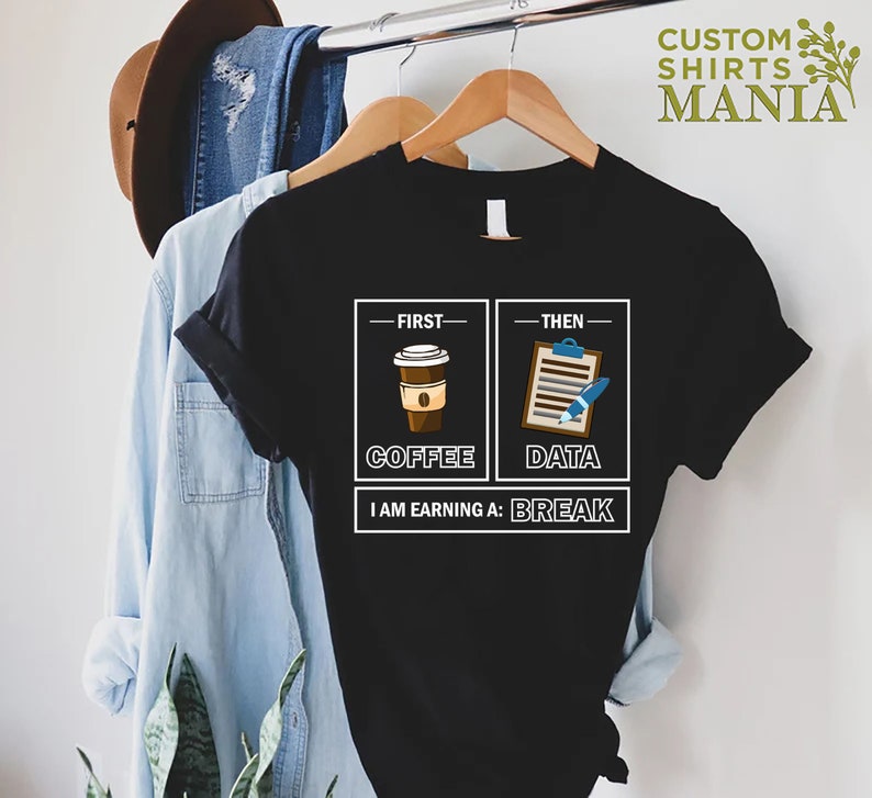 May include: Black t-shirt with a white graphic design. The graphic shows two boxes, one with a cup of coffee and the text "First - Coffee" and the other with a clipboard and pen and the text "Then - Data". Below the boxes is the text "I am earning a: Break".