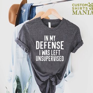 May include: A dark gray t-shirt with white text that reads "In my defense I was left unsupervised".