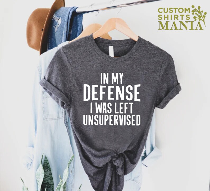 May include: A dark gray t-shirt with white text that reads "In my defense I was left unsupervised".