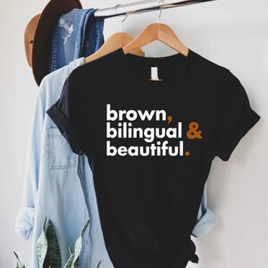 Puede incluir: Camiseta negra con texto blanco que dice "brown, bilingual & beautiful." El texto est&aacute; en negrita y la ampersand es de color naranja.