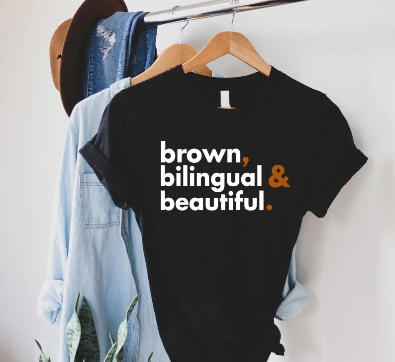 Puede incluir: Camiseta negra con texto blanco que dice "brown, bilingual & beautiful." El texto est&aacute; en negrita y la ampersand es de color naranja.