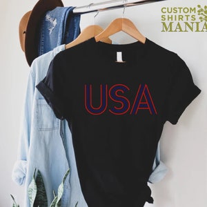 Może przedstawiać: Czarny T-shirt z czerwono-niebieskim nadrukiem "USA". Koszulka wisi na drewnianym wieszaku.