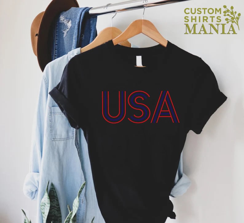 Może przedstawiać: Czarny T-shirt z czerwono-niebieskim nadrukiem "USA". Koszulka wisi na drewnianym wieszaku.