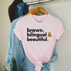 Puede incluir: Camiseta rosa con el texto "brown, bilingual & beautiful." en negro y naranja.