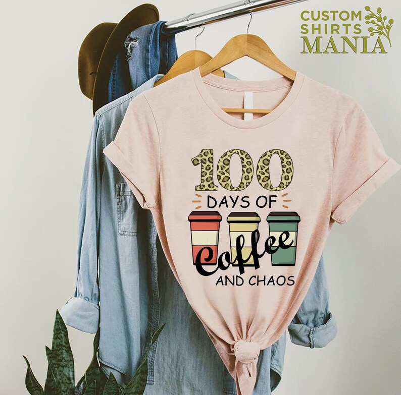 Pu&ograve; includere: Maglietta rosa con un design grafico di tre tazze di caff&egrave; e il testo "100 giorni di caff&egrave; e caos". Il numero 100 &egrave; decorato con uno stampa leopardato.