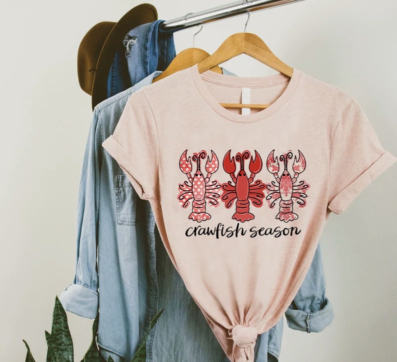 May include: A light pink t-shirt with three red crawfish illustrations. The crawfish have different patterns, including polka dots and floral designs. The words "crawfish season" are written in black script below the crawfish.