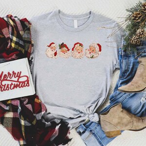 May include: A gray t-shirt with a vintage-style print of four Santa Claus faces. The shirt is tied in a knot at the bottom. The shirt is surrounded by a plaid scarf, a pair of jeans, and a pair of brown boots. A white sign with the words "Merry Christmas" in red letters is also visible.