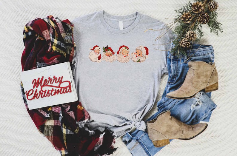 May include: A gray t-shirt with a vintage-style print of four Santa Claus faces. The shirt is tied in a knot at the bottom. The shirt is surrounded by a plaid scarf, a pair of jeans, and a pair of brown boots. A white sign with the words "Merry Christmas" in red letters is also visible.