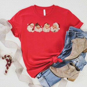 May include: Red t-shirt with a graphic of five vintage Santa Claus faces. The shirt is tied in a knot and is laying on a white surface with a pair of blue jeans and brown ankle boots.