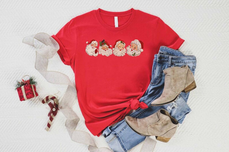 May include: Red t-shirt with a graphic of five vintage Santa Claus faces. The shirt is tied in a knot and is laying on a white surface with a pair of blue jeans and brown ankle boots.