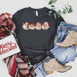 May include: A dark gray t-shirt with a vintage-style print of four Santa Claus faces. The shirt is tied in a knot and is laying on a white surface with a plaid blanket, a pair of jeans, and two brown ankle boots.